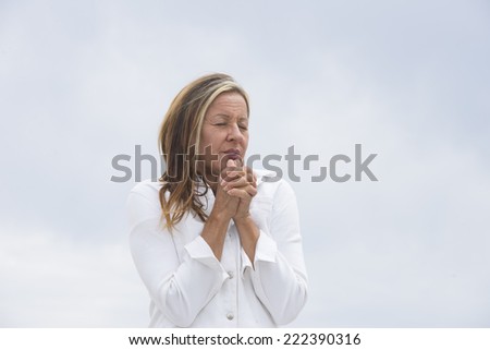 Portrait attractive mature woman in white outdoor, with folded praying, begging hands and closed eyes, bright background and copy space.