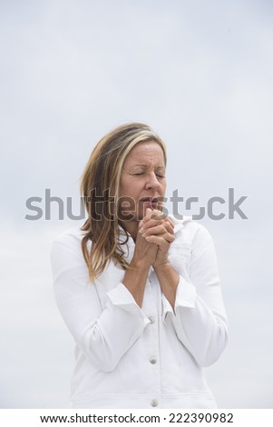 Portrait attractive mature woman with closed eyes in white outdoor, with folded praying, begging hands and closed eyes, bright background and copy space.