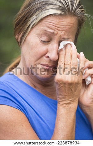 Portrait crying mature woman with sad unhappy emotional facial expression, in grief or suffering painful depression, with tissue in hands, outdoor blurred background.