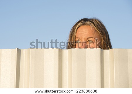 Portrait of woman watching, observing hiding behind neighbor metal fence outdoor, with blue sky as background and copy space