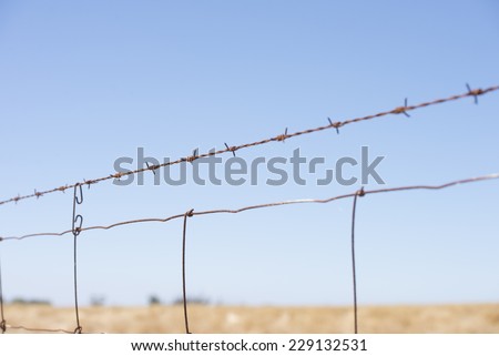 Rusty Barb wire fence along paddock or farm land in rural agricultural country, with farm land and blue sky as blurred background and copy space.