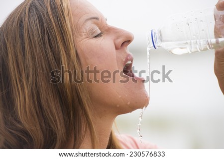 Portrait attractive mature woman drinking water as refreshment after sport activity outdoor, spills and drops over face with bright background and copy space.