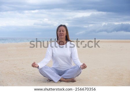 Portrait mature woman meditating at beach with closed eyes concentrated and relaxed, with ocean and cloudy sky as blurred background and copy space.