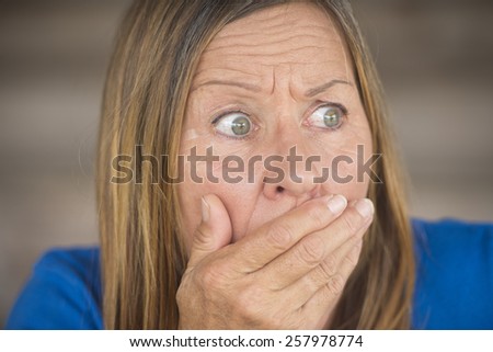 Portrait attractive mature woman with shocked, anxious, fearful facial expression, covering mouth with hand, blurred background.