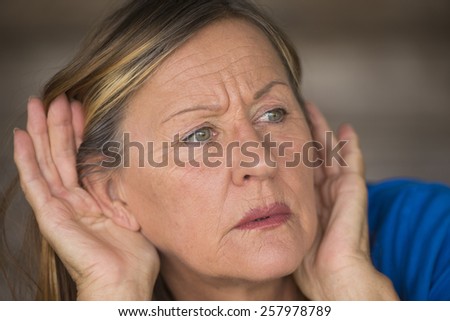 Portrait attractive mature woman with hands at ears listening with curious, interested and worried facial expression to loud noise or sound, blurred background.