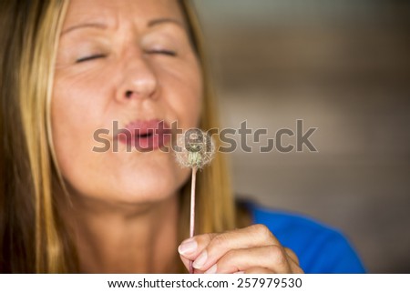 Portrait of attractive mature woman in blurred background blowing dandelion flower in hand, happy, friendly, closed eyes, copy space.