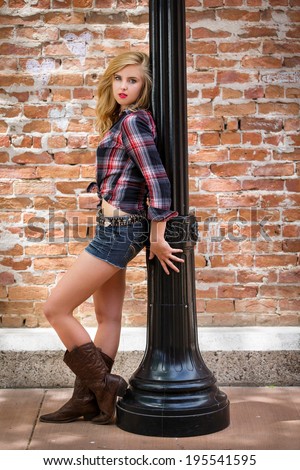 Cute and young blond girl posing in boots and flannel shirt next to a post