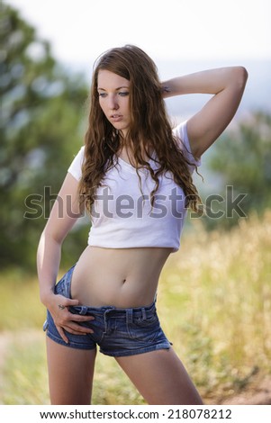 Young woman with red hair posing with one arm over her head and wearing low rise jeans shorts