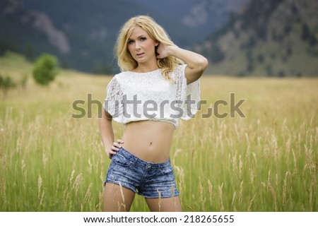 Joyful young blond lady with a smile posing in short white top and denim jeans shorts with grassy field in the background