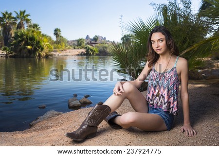 Young brunette girl posing and sitting next to a lake with cowboy boots and denim shorts