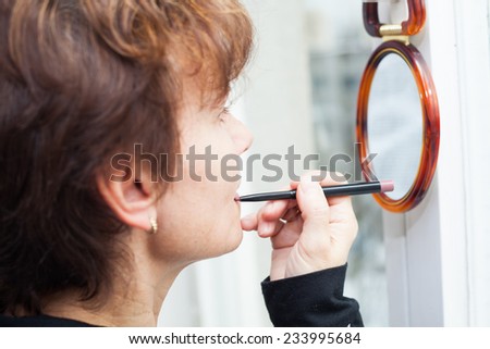 Middle-aged woman applying lip pencil and looking in mirror.
