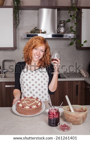 Happy redhead woman in love showing chocolate cake and holding glass of wine in the kitchen at home.