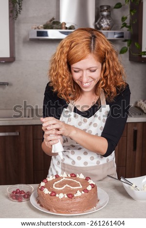 Happy redhead woman decorating chocolate cake with a love heart, in the kitchen at home.