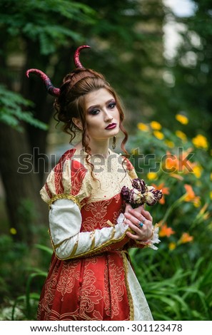 Beautiful girl with a demonic horns in medieval red dress on a background of nature with dry roses in their hands