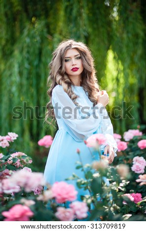 Beautiful girl with long bushy curly hair in a gentle mint-colored long dress walking on a beautiful green garden with blooming red and pink roses