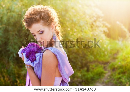 Beautiful young woman in a purple dress with red hair and a bouquet of purple asters in the park in the evening