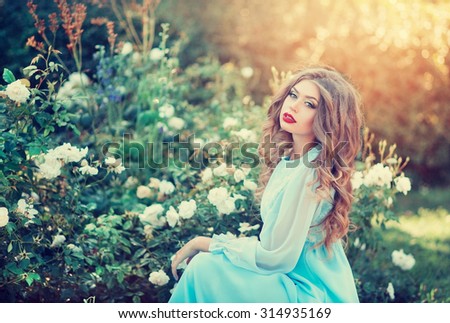 Lovely young woman in a long dress the color of mint with long curly hair in a summer garden with blooming roses