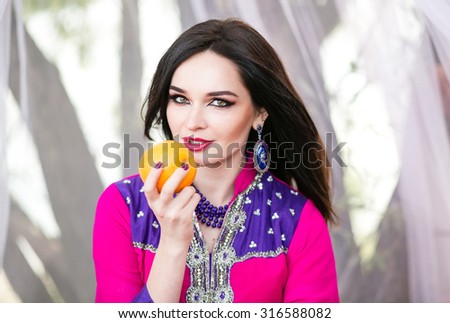 Beautiful dark-haired girl of Arab appearance, dressed in traditional bright oriental costume sitting in a white tent with an orange in the hands orange, closeup