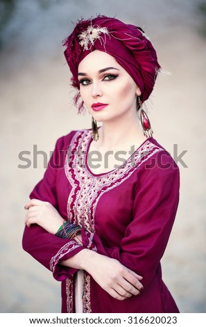 Portrait of a beautiful woman with make-up in the Arab east suit and turban, sitting in the desert, closeup
