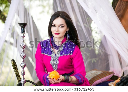 Beautiful dark-haired girl of Arab appearance, dressed in traditional bright oriental costume sitting in a white tent with an orange in the hands orange