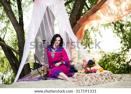 Beautiful young dark-haired woman in a colorful Arabic garb sitting under a white tent, smoke hookah, oriental style
