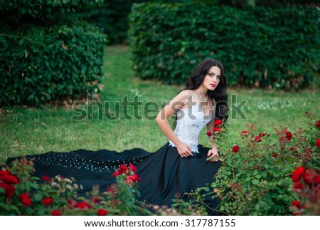 Beautiful dark-haired girl who looked like a princess in a long evening dress in the park with stunning beautiful red roses at sunset