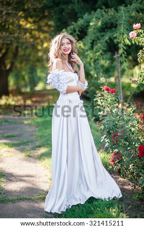 Beautiful girl bride in a long white satin dress walks on summer garden with flowers