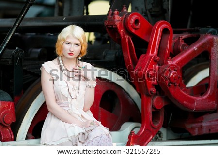 Beautiful feminine blonde girl in vintage pink dress sits near a large massive iron wheels of an old steam locomotive