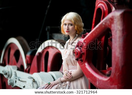 Beautiful feminine blonde girl in vintage pink dress sits near a large massive iron wheels of an old steam locomotive