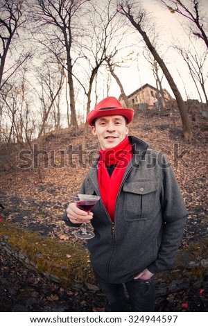 A nice young man in elegant black coat and red scarf and hat in autumn park with a glass of red wine
