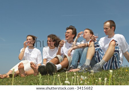 Five nice young people in white vests sit on a grass and drink water from plastic glasses