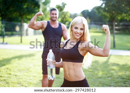 Fit couple in nature with bottle of water, blonde woman showing biceps, healthy lifestyle