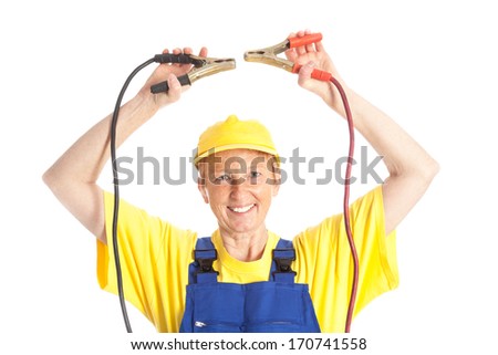 elder female mechanic in as blue overall bib and with a yellow cap holding a red and black battery booster cable for making a short in front of a white background