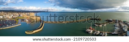 Wide aerial panorama of Carrickfergus near Belfast with medieval Norman castle, harbor, marina, yachts, breakwater, boat ramp, Belfast Laugh, parking lot. Sunset light and strom clouds in winter.