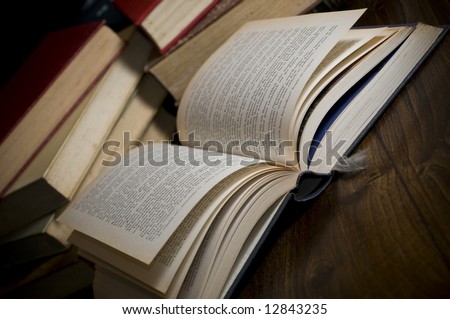 high resolution picture of an old book over a wooden table. Piles of other books are on the background and the light cast on the open pages create a moody atmosphere.