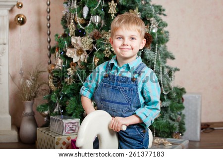 Little boy in denim overalls riding a wooden horse near the Christmas tree