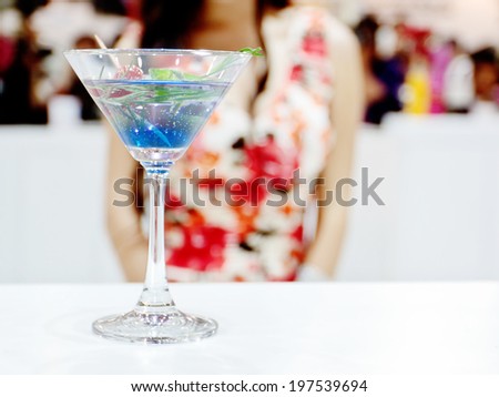 A glass of a blue cocktail on white table with a sexy girl on a background