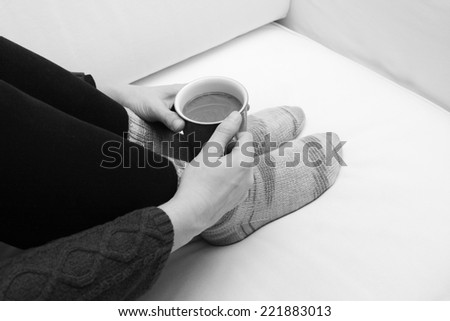 Woman\'s hands holding a mug of hot coffee or tea, curled up on the sofa, wearing cosy hand-knitted socks - monochrome processing