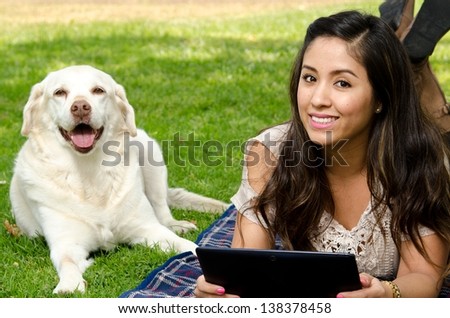 a smiling Hispanic woman in the park with her pet dog and holding a tablet.