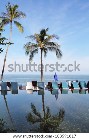 Pool on a beach of the Thai gulf. The palm tree and beach armchairs are reflected in a smooth water surface