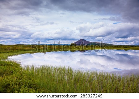 In the smooth water of cold lake reflects cloudy sky. Summer Iceland. Small lake surrounded by green meadow