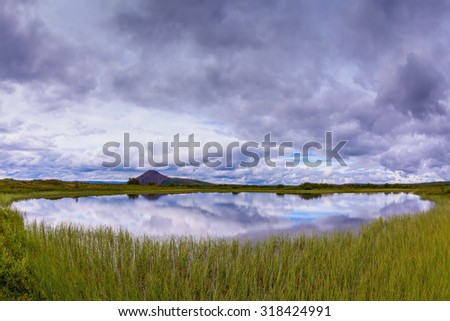 Small lake surrounded by green meadow. In the smooth water of cold lake reflects cloudy sky. Summer Iceland
