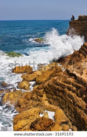 The fisherman sits with a fishing tackle at the sea on top  an ancient stone wall