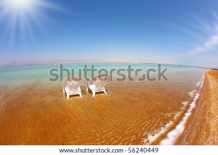 Two white beach chairs stood side by side in the clear water. The sun shines on the beach therapeutic