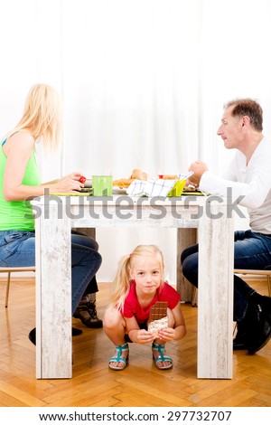 Girl eating chocolate beneath table while the family is having breakfast
