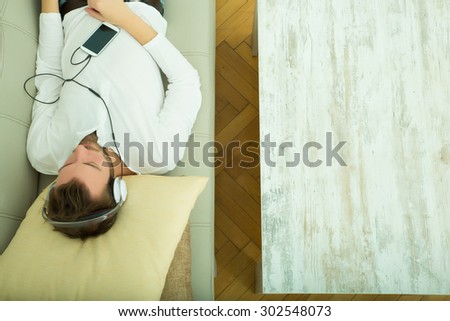 A young man lying on the sofa listening to music with some headphones and a smartphone.