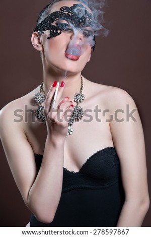 Fashion photo of gorgeous woman with dark hair and blue eyes, with lace mask on her face,posing in dark studio. Sexy Woman. Smoking Woman.