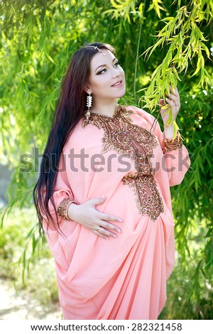 Beautiful pregnant woman in the park. Indian girl in dress outdoor. Pregnant woman feeling the baby.