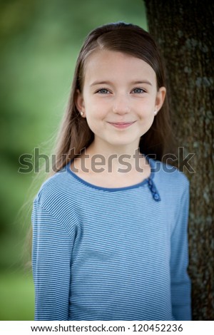 Young Caucasian girl with brown hair, blue eyes, and blue shirt standing next to a tree