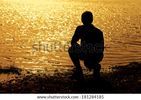 Silhouette of teenage boy kneeling and pondering in front of sunset over water